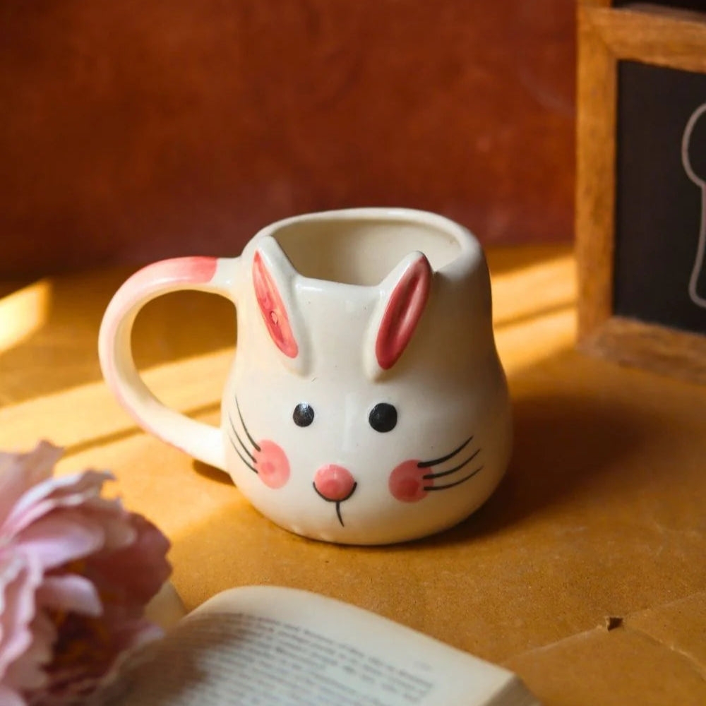 Ceramic mug shaped like a rabbit with a pink flower and open book on a wooden surface.