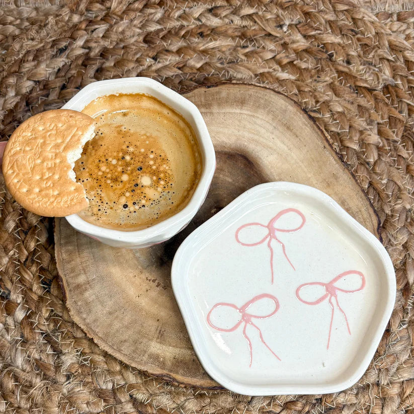 Cup of coffee with a cookie on a wooden coaster, accompanied by a small white dish with pink bow designs.