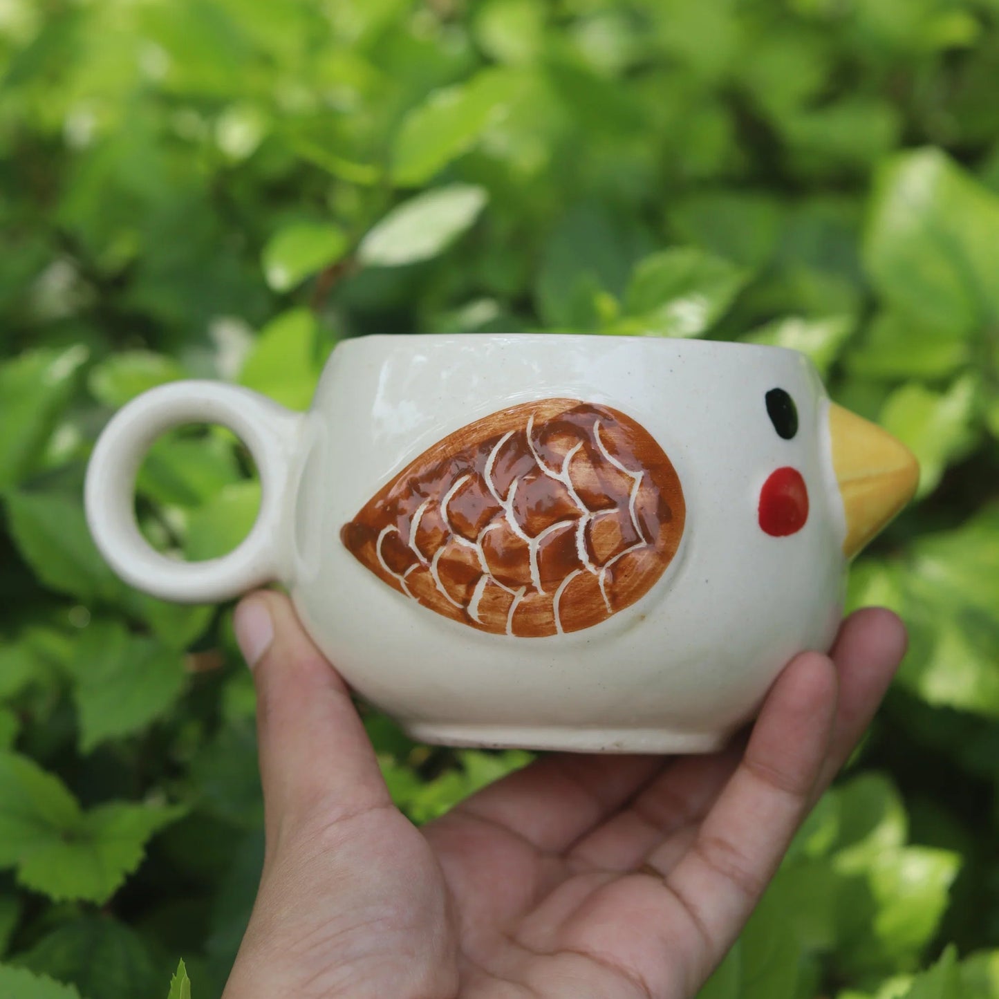 Hand holding a ceramic chicken-shaped mug against a green leafy background