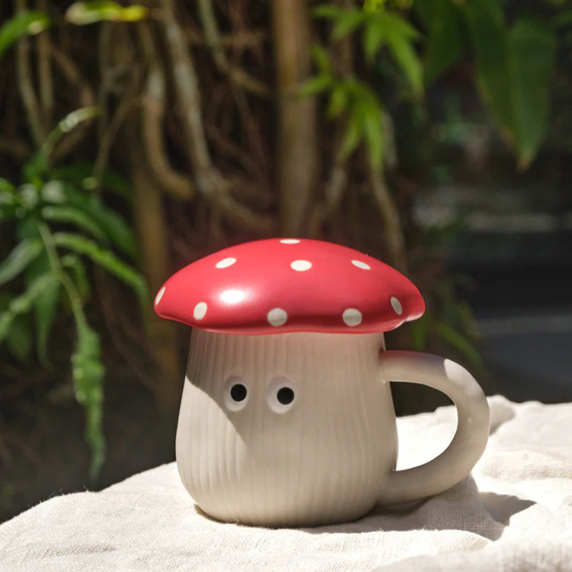 Mug with a red mushroom lid on a white surface with greenery in the background