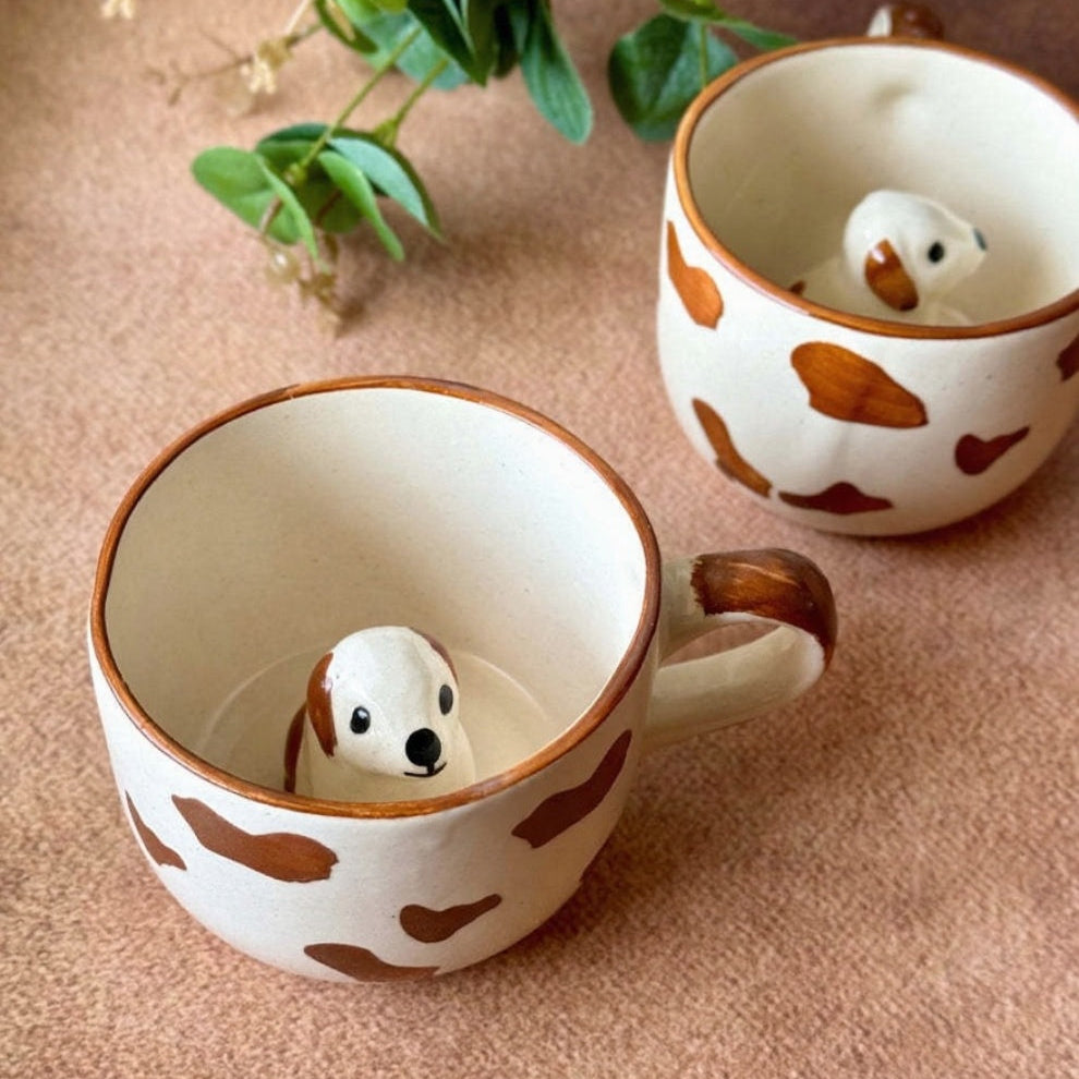 Two ceramic mugs with dog-shaped figures inside, on a beige surface with a plant in the background.