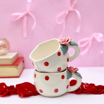 Two stacked ceramic mugs with red polka dots and floral handles on a pink background with decorative bows.