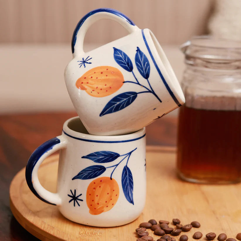 Two ceramic mugs with orange and leaf design on a wooden surface with coffee beans and a glass of coffee.