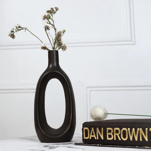 Black vase with dried flowers on a white surface next to a book titled 'Dan Brown'.