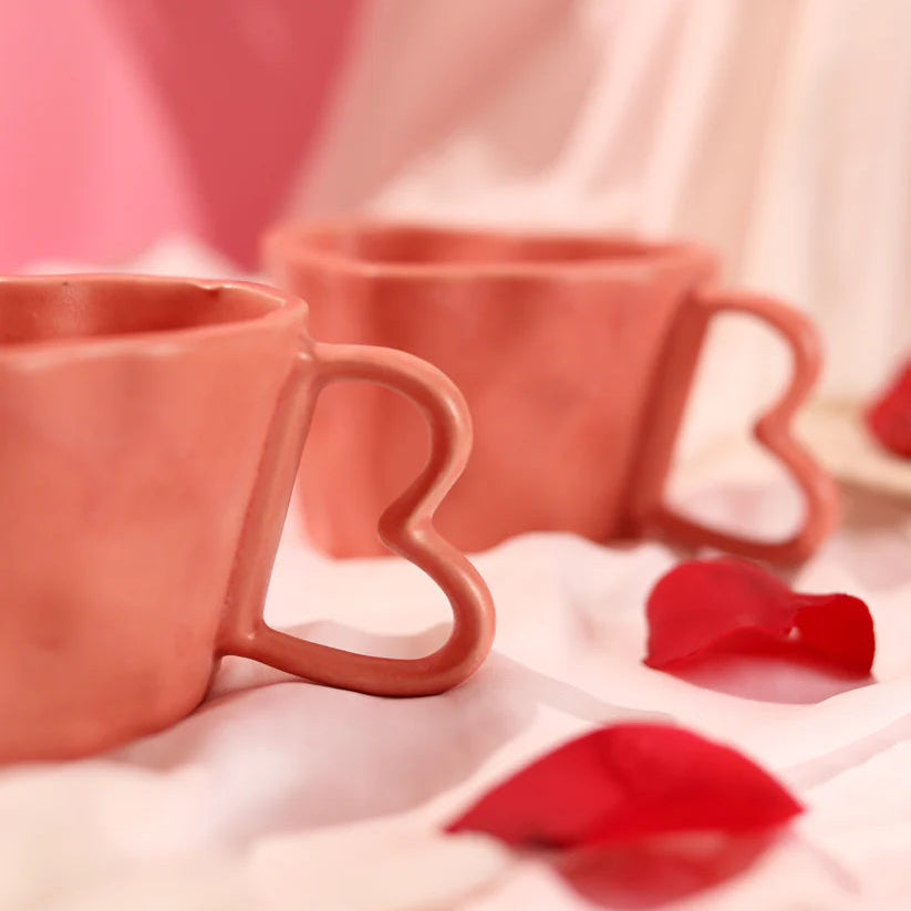 Two heart-shaped pink mugs on a soft pink background with red heart-shaped objects.