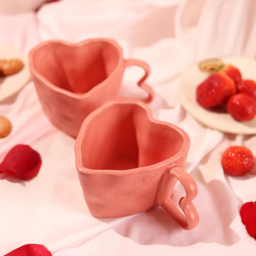 Two heart-shaped pink mugs on a soft pink fabric background with strawberries.