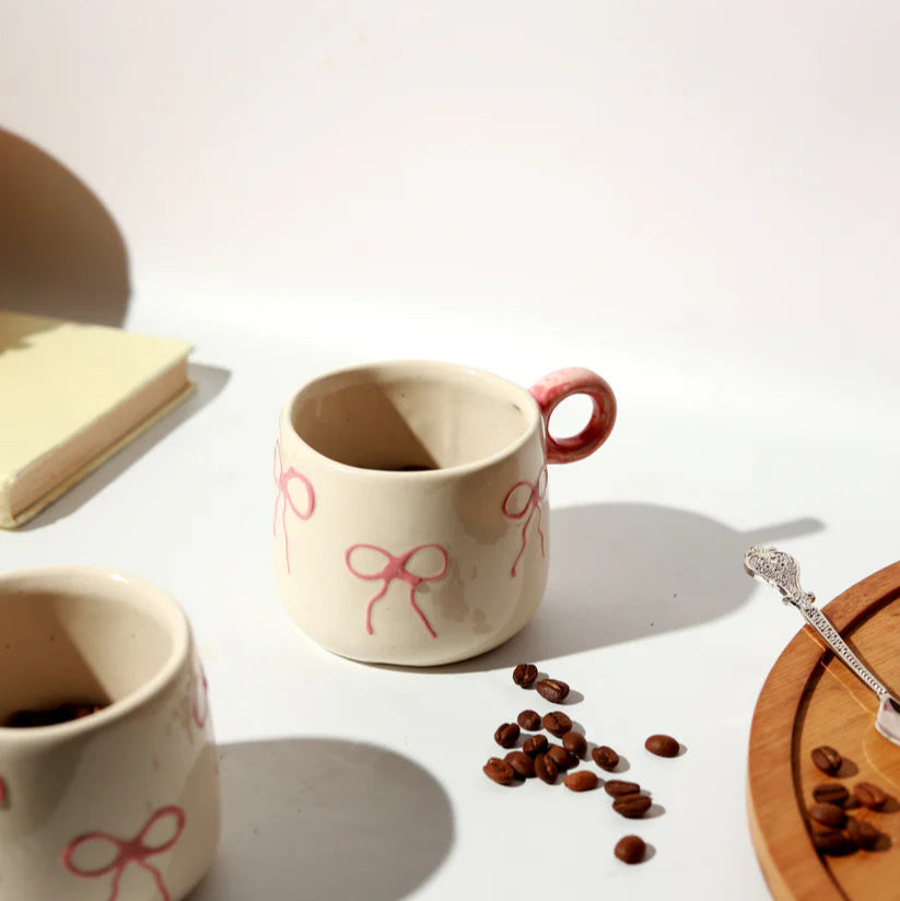 Two ceramic mugs with red bow designs, coffee beans, and a wooden coaster on a white surface.