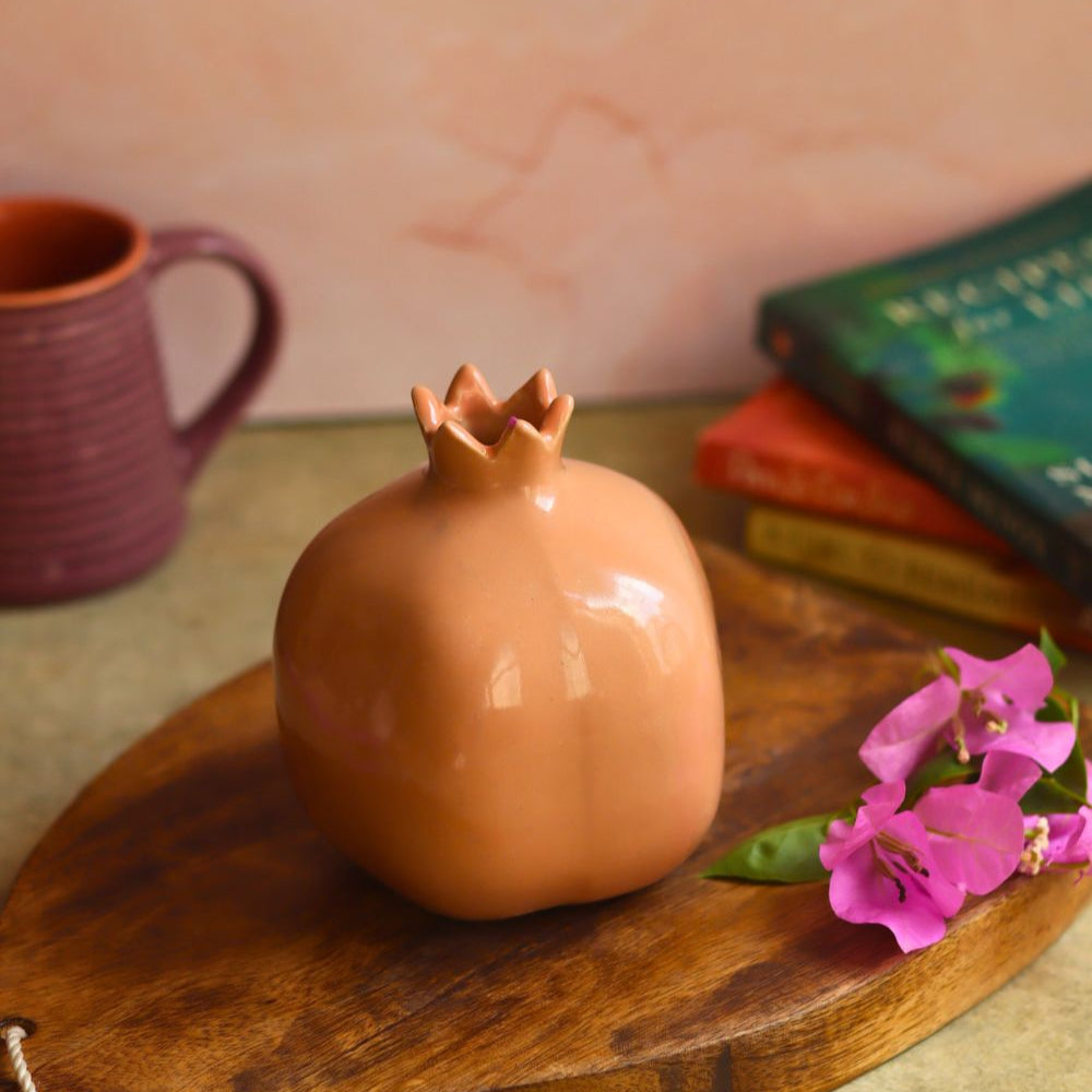 Ceramic pomegranate-shaped object on a wooden board with a pink flower and books in the background.