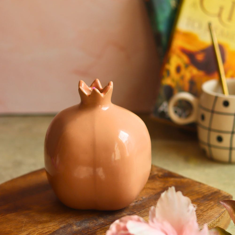 Ceramic pomegranate-shaped object on a wooden surface with books and a mug in the background.