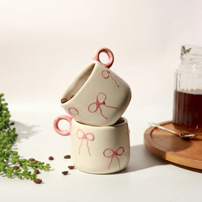 Two ceramic mugs with bow designs stacked on a light surface, accompanied by a glass of tea and a wooden coaster.