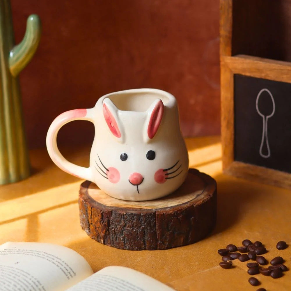 Ceramic mug shaped like a rabbit with a wooden base on a table with coffee beans and an open book.