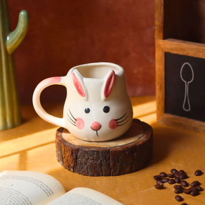 Ceramic mug shaped like a rabbit with a wooden base on a table with coffee beans and an open book.