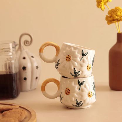 Two stacked ceramic mugs with floral patterns on a table with a glass of coffee and vase in the background.
