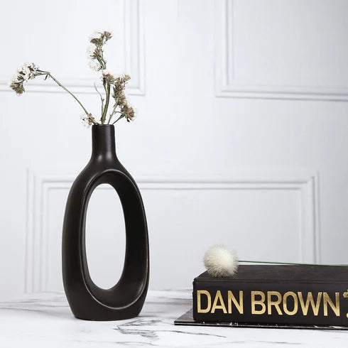 Black vase with dried flowers on a surface with 'Dan Brown' book, against a white wall.