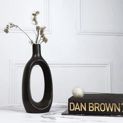 Black vase with dried flowers on a surface with 'Dan Brown' book, against a white wall.