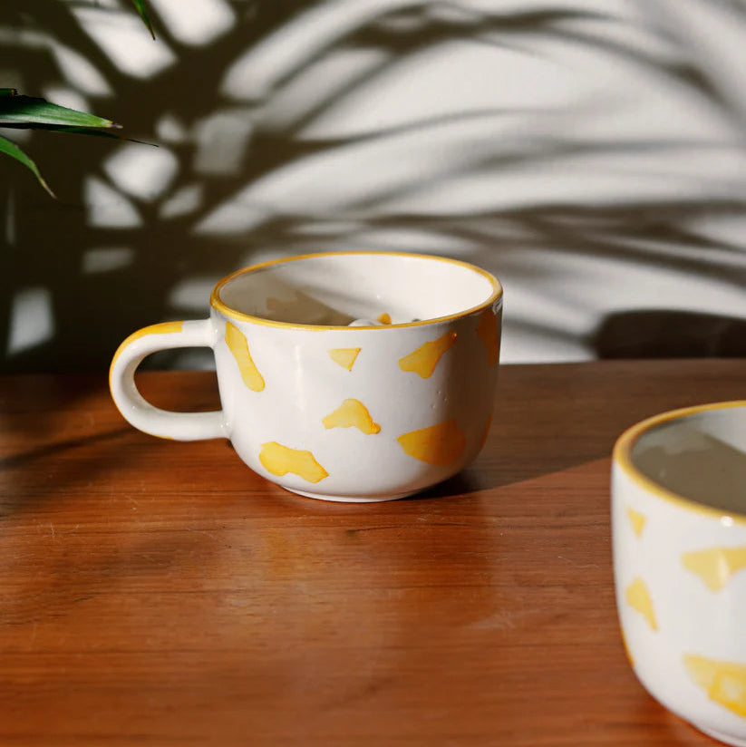 Two ceramic mugs with yellow patterns on a wooden surface, casting shadows.