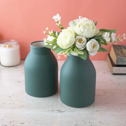 Two green vases with a bouquet of white flowers on a wooden surface against a pink wall.