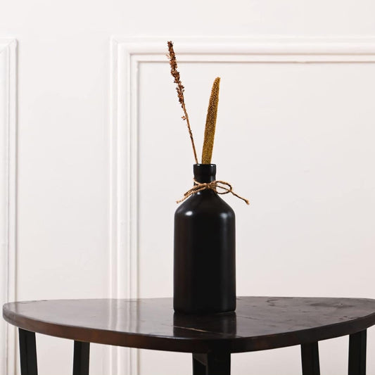 Black vase with dried plants on a wooden table against a white wall.