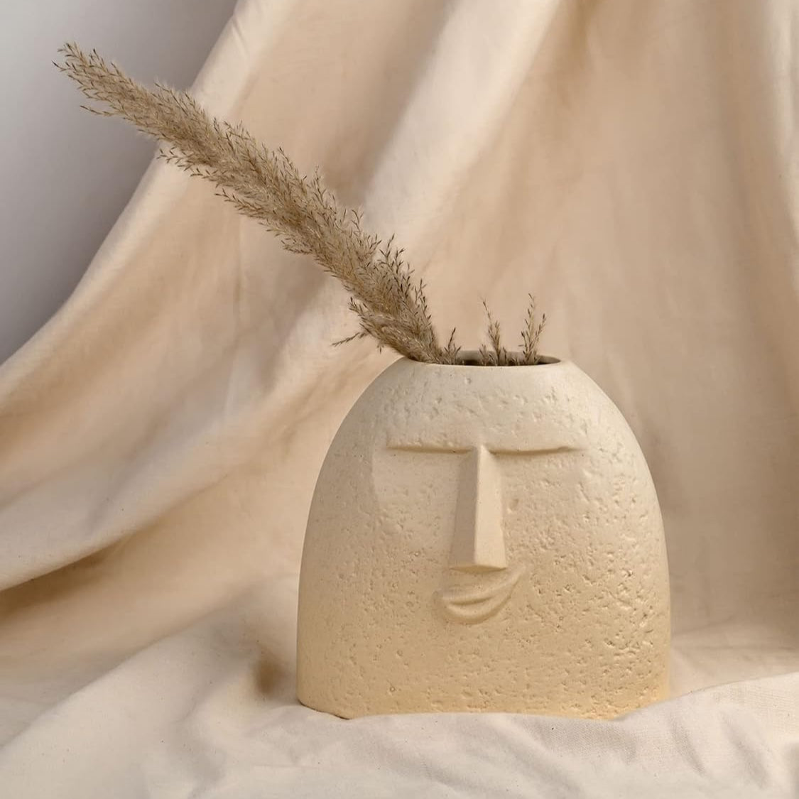 Beige textured vase with dried plant on a soft beige fabric background