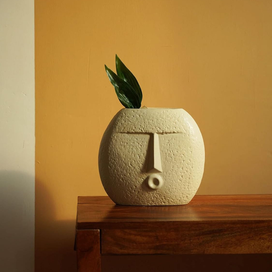 Ceramic vase shaped like a face with a plant on a wooden surface against an orange wall.