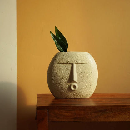 Ceramic vase shaped like a face with a plant on a wooden surface against an orange wall.