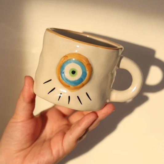 Ceramic mug with eye design held by a hand against a neutral background