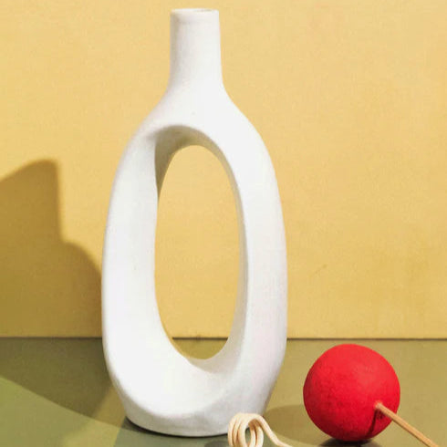 White ceramic vase with a red ball and sticks on a reflective surface against a yellow background