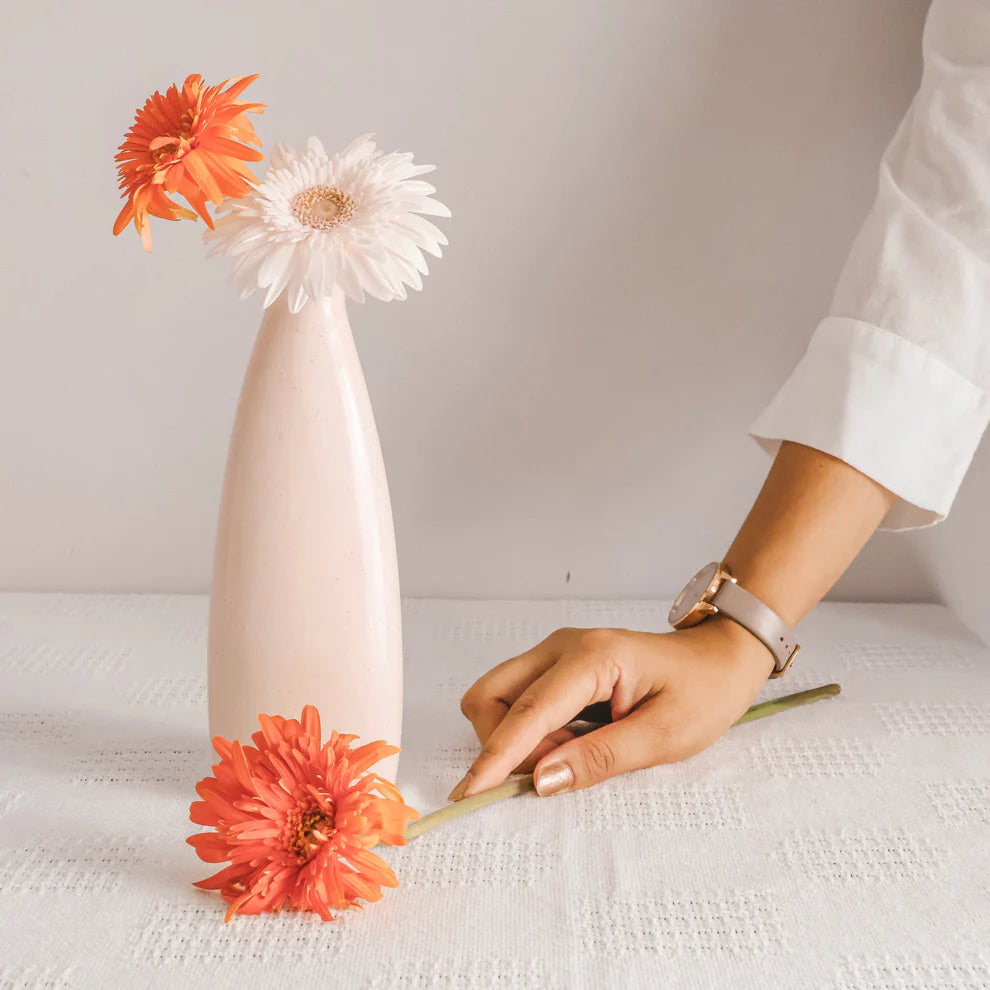 Person arranging flowers around a tall, thin vase on a white surface.