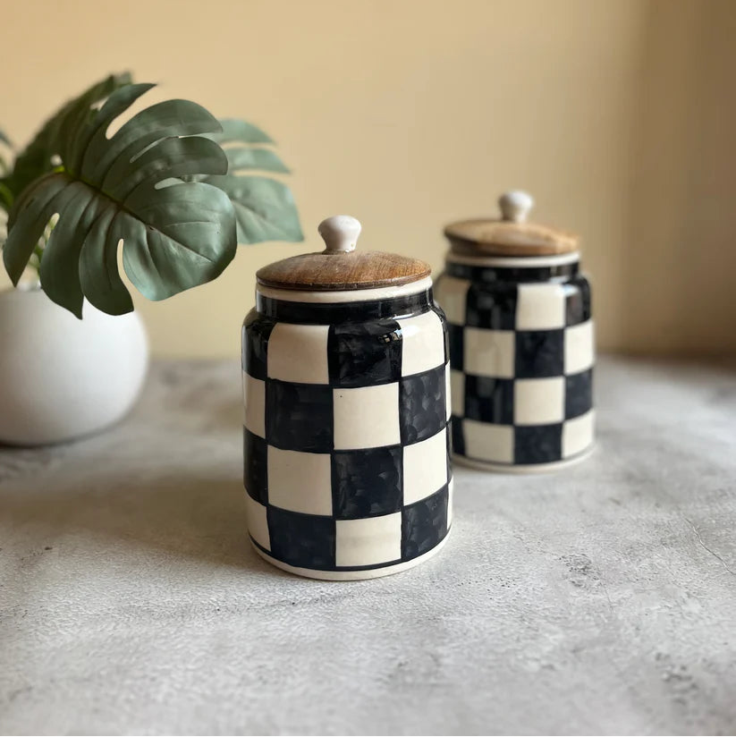 Two black and white checkered jars with wooden lids on a gray surface with a plant in the background.