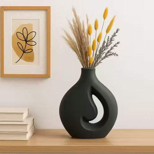 Black decorative vase with dried plants on a wooden shelf, framed artwork in the background.