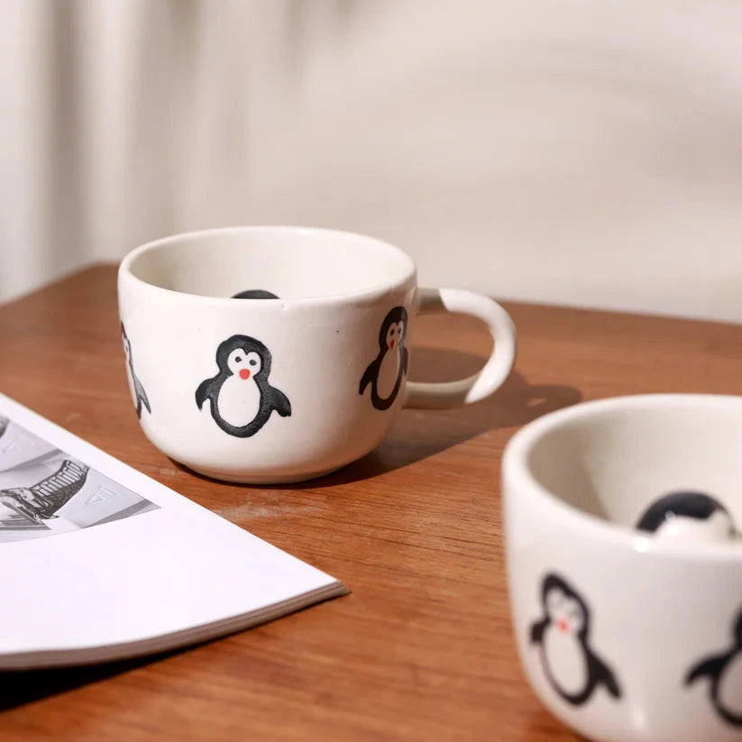 Two white mugs with penguin designs on a wooden surface.