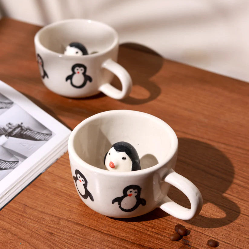 Two white mugs with penguin designs on a wooden surface with coffee beans.