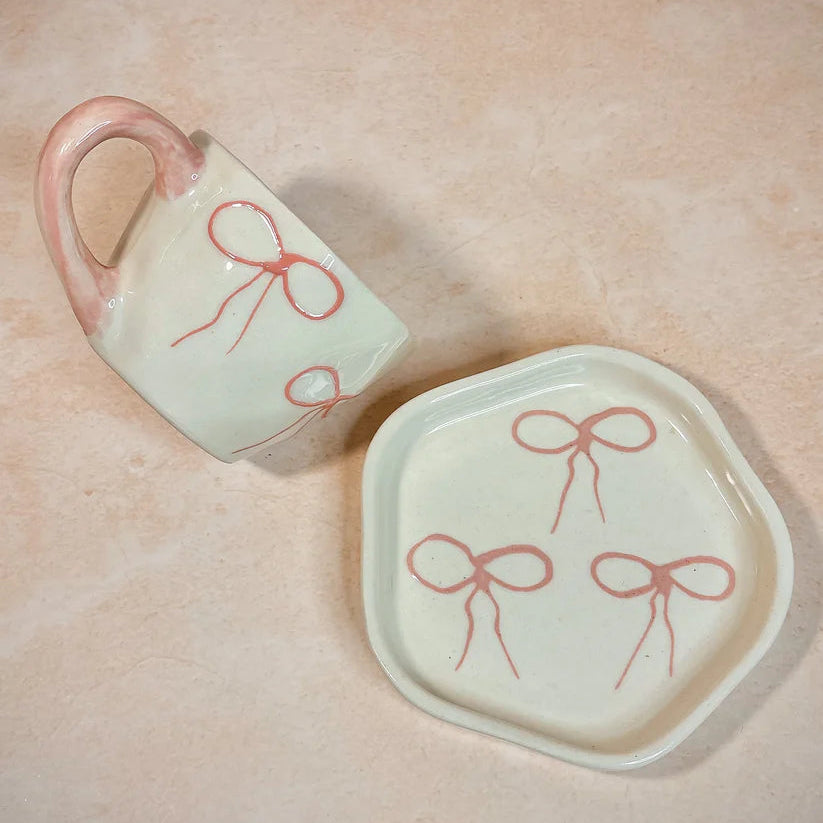 Ceramic mug and plate with red bow designs on a beige background