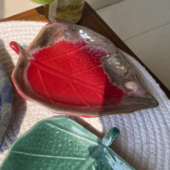 Three leaf-shaped ceramic bowls in blue, red, and green on a textured surface.