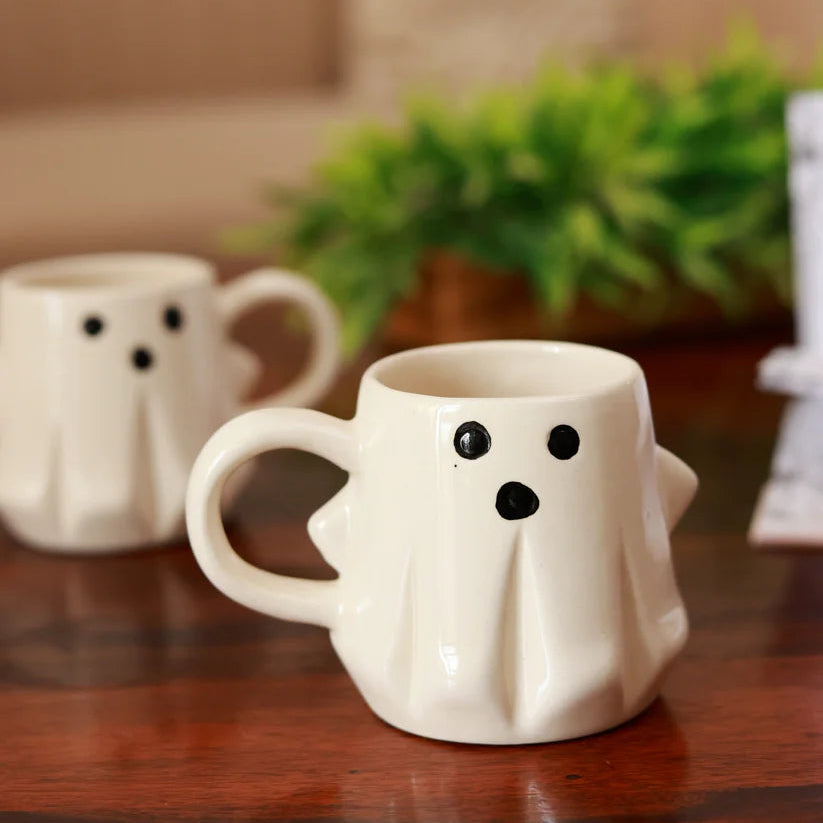 Two ghost-shaped ceramic mugs on a wooden surface with a blurred background