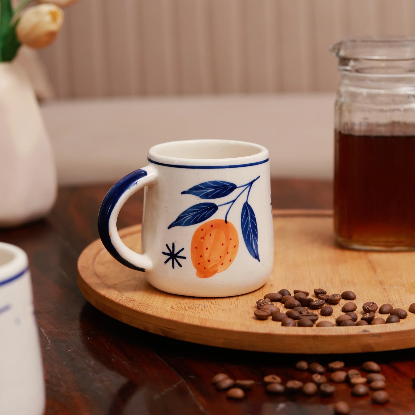 White mug with blue handle and orange design on a wooden coaster with coffee beans and a jar of coffee in the background.