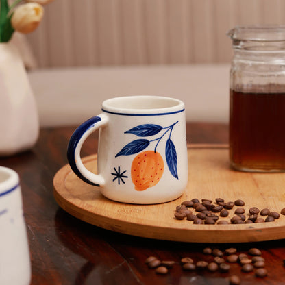 White mug with blue handle and orange design on a wooden coaster with coffee beans and a jar of coffee in the background.