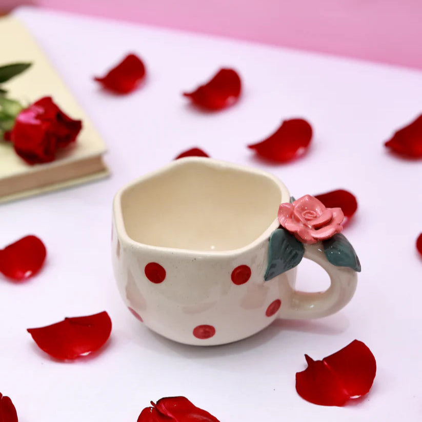 Ceramic mug with red polka dots and a pink rose on a white surface with red rose petals.