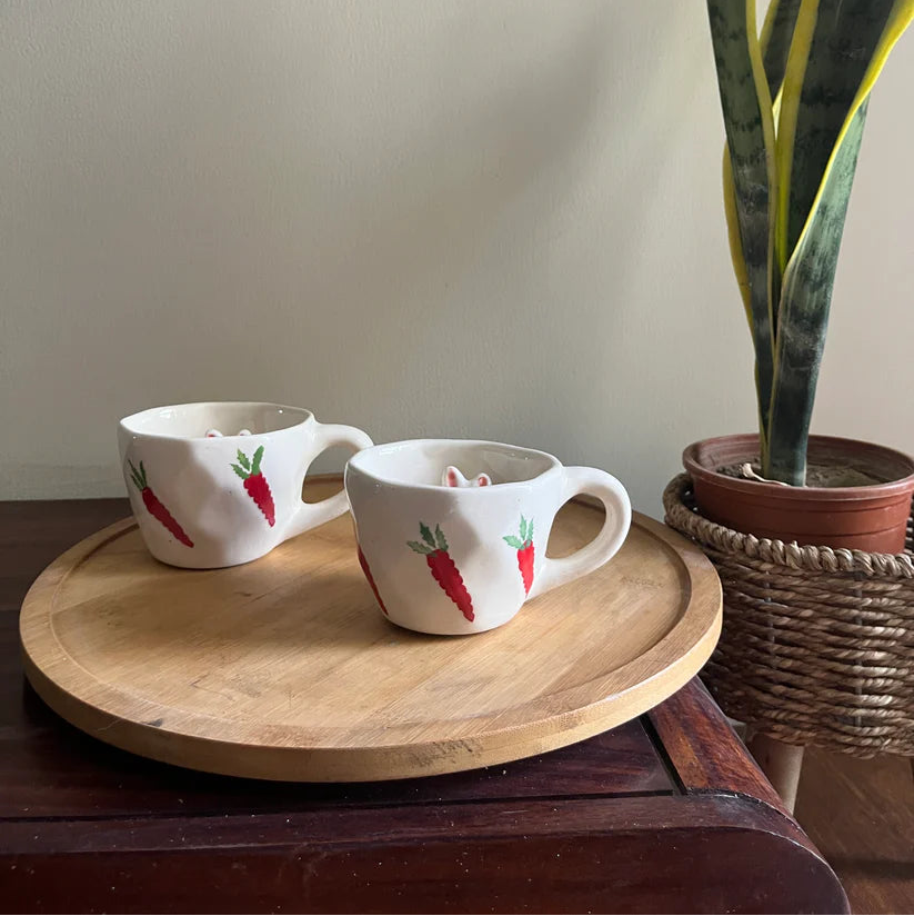 Two mugs with carrot design on a wooden tray next to a potted plant.