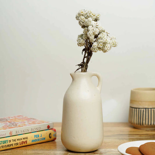 White ceramic pitcher with dried flowers on a wooden surface with books and cookies.