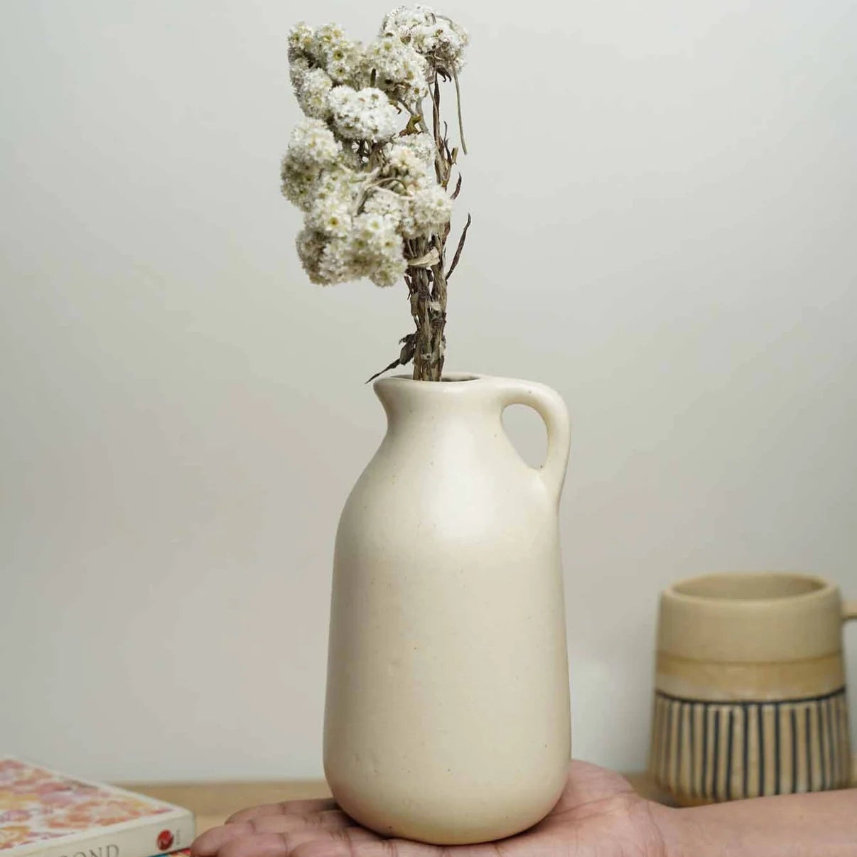 Person holding a beige ceramic vase with dried flowers on a wooden surface.