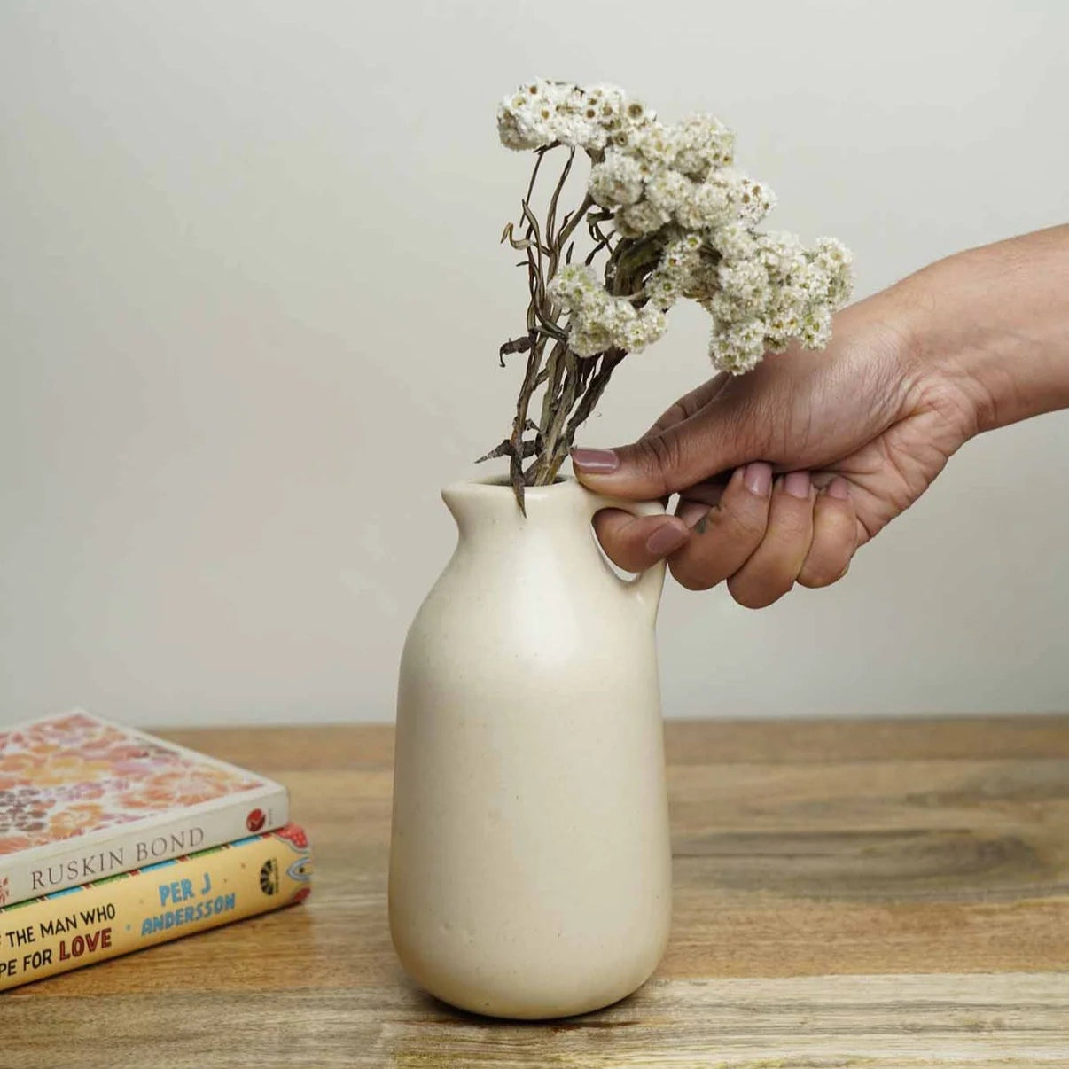 Hand placing dried flowers into a small beige vase on a wooden table.