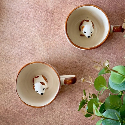Two ceramic mugs with dog-shaped handles on a pink surface, accompanied by a small plant.