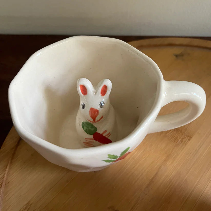 Small ceramic rabbit figurine with a carrot inside a white mug on a wooden surface.