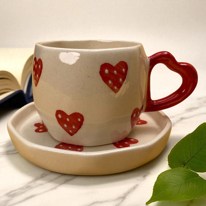 Ceramic mug with red heart designs on a matching saucer, placed on a marble surface with a green leaf.