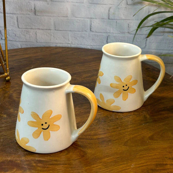 Two ceramic mugs with yellow floral designs on a wooden table against a white brick wall.