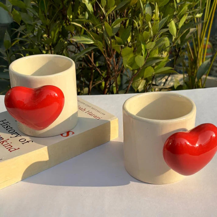 Two ceramic cups with red heart shapes on a white surface with greenery in the background.