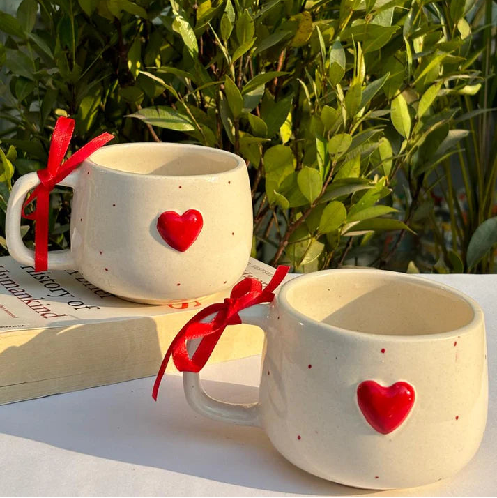 Two ceramic mugs with red heart designs on a white surface with greenery in the background.