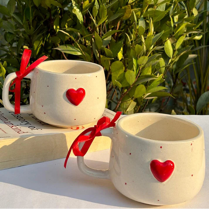 Two ceramic mugs with red heart designs on a white surface with greenery in the background.