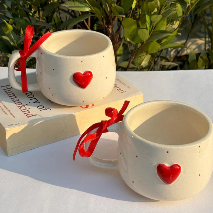 Two ceramic mugs with red heart designs on a white surface with greenery in the background.